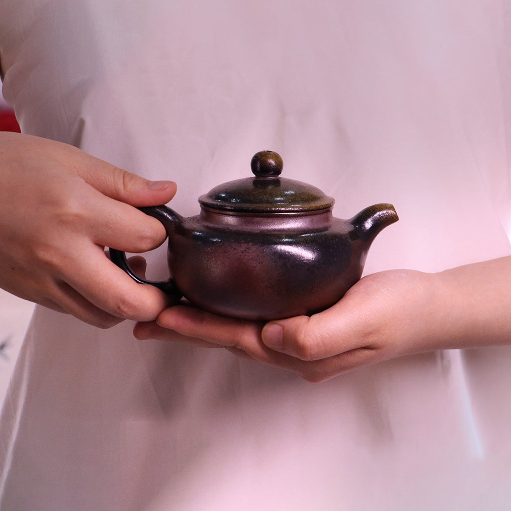 Chinese Handcrafted Fanggu Teapot in deep purple ceramic held by hands with soft pink background