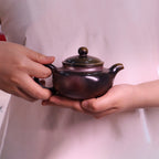 Chinese Handcrafted Fanggu Teapot in deep purple ceramic held by hands with soft pink background
